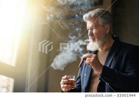 Handsome brutal middle aged man with beard looking away while holding cigar and glass of whisky, standing in loft interior 83001944