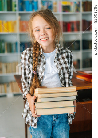 Vertical portrait of smiling elementary child school girl holding stack of books in library at school, looking at camera. 83002786