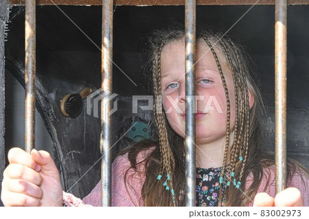 Sad little girl behind iron bars. Little girl behind the bars. Violence concept Sad little girl behind iron bars. Little girl behind the bars. Violence concept 83002973