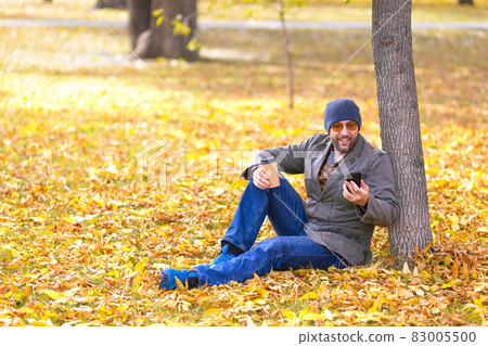Middle aged man during online video call in an autumn park - he is sitting under the tree on fallen leaves and looking at his smartphone. 83005500