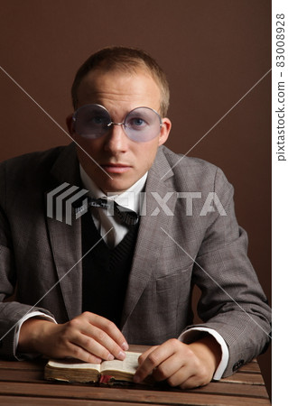 A handsome, confident businessman in a suit and round glasses, sitting isolated on a brown background, reading a book. A man's thoughtful, surprised face. High quality photo 83008928