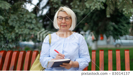 Portrait of attractive adult female teacher 35-40 years old in vision glasses looks into the camrera in the park. Woman writing in a notebook. Portrait of attractive adult female teacher 35-40 years old in vision glasses looks into the camrera in the park. Woman writing in a notebook. 83014137