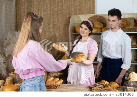 A young, attractive saleswoman and a handsome baker show their bread to a teenage client so that she can choose and buy one. 83014306