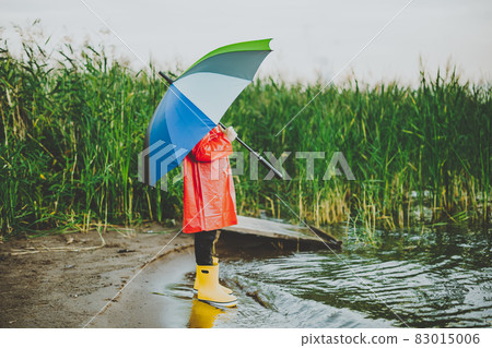 Boy in a red raincoat and yellow rubber boots stands at river bank and holding rainbow umbrella. School kid standing still near autumn lake. Child wearing waterproof clothes at shoreside 83015006