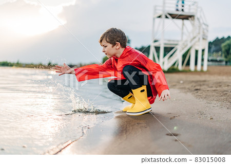 Boy in a red raincoat and yellow rubber boots playing with water at the beach. School kid in a waterproof coat touching water at sea. Child having fun with waves at the shore 83015008