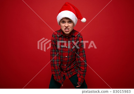 Funny preadolescent boy in plaid shirt and Santa Claus hat grimacing looking at camera posing against red colored background with copy space for Christmas and New Year ad 83015983
