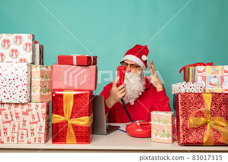Santa Claus wearing hat work in his oficce with presents, preparing for holidays. Santa Claus wearing hat work in his oficce with presents, preparing for holidays. 83017315
