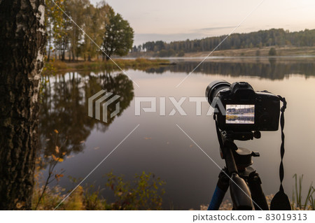 black digital camera on tripod shooting early foggy morning landscape at autumn lake with selective focus 83019313