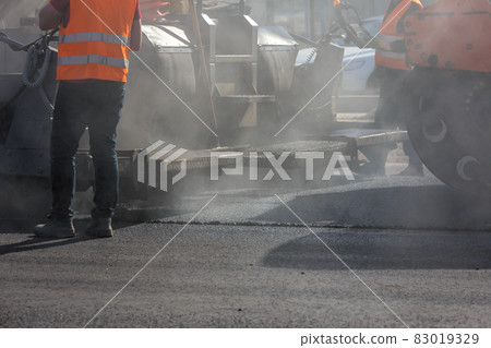 men working with asphalting paver machine during road street repairing works at day light with smoke and steam in the air 83019329