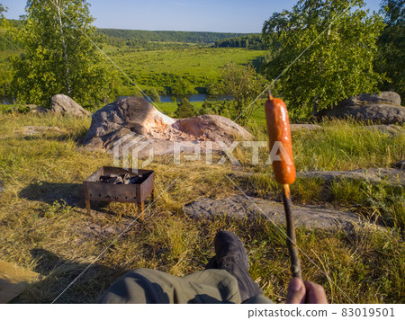 fried sausage on a stick in front of portable brazier at wild riverside camping - gonzo view of nature recreation 83019501