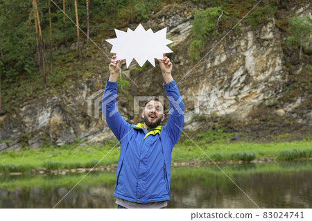 Smiling young man with beard in jacket holding blank speech bubble above your head on background of natural landscape Smiling young man with beard in jacket holding blank speech bubble above your head on background of natural landscape 83024741