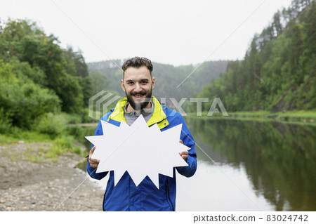 Smiling young man with beard in jacket holds white star board in front of him against background of natural landscape Smiling young man with beard in jacket holds white star board in front of him against background of natural landscape 83024742
