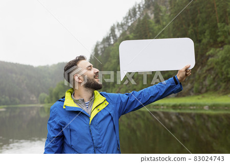 Young man with beard in jacket holds empty blank board against background of landscape of river and forest. Young man with beard in jacket holds empty blank board against background of landscape of river and forest. 83024743