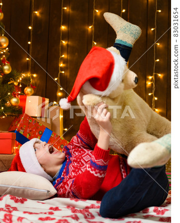 Child girl posing and having fun in new year or christmas decoration. Holiday lights and gifts, Christmas tree decorated with toys. She's wearing a red sweater and a Santa helper hat 83031654