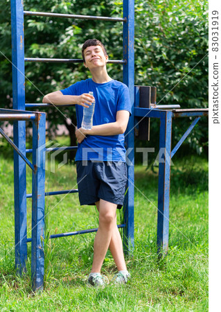 teenage boy exercising outdoors, sports ground in the yard, he stands near the horizontal bar, opens a bottle of water and drinks, healthy lifestyle teenage boy exercising outdoors, sports ground in the yard, he stands near the horizontal bar, opens a bottle of water and drinks, healthy lifestyle 83031919