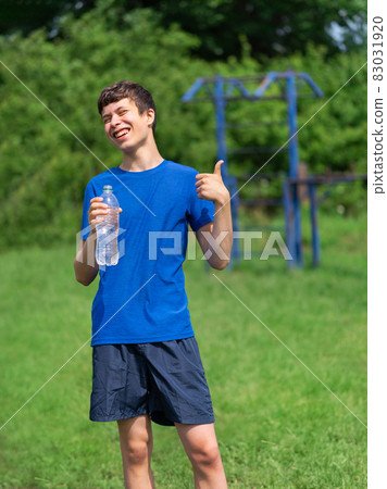 teenage boy exercising outdoors, sports ground in the yard, he opens a bottle of water and drinks, healthy lifestyle 83031920