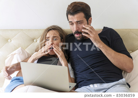 Scared young couple sitting under blanket on the bed with laptop while watching horror movie at tv Scared young couple sitting under blanket on the bed with laptop while watching horror movie at tv 83036859