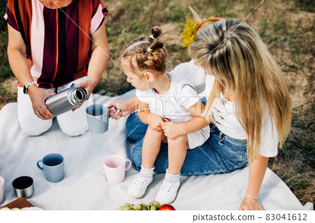 Close-up of a grandmother's hand pouring tea from a thermos to her granddaughter, sitting in her mother's arms at a picnic 83041612