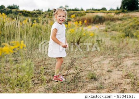 A happy little girl in a white dress stands on a field with yellow flowers and smiles on a warm day 83041613
