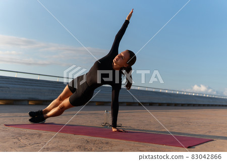 Female athlete doing side plank exercise with raised arm on mat during fitness training on embankment against cloudy blue sky. 83042866