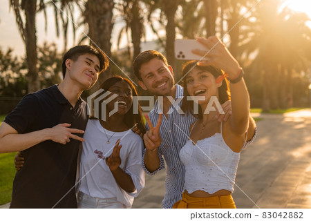 Company of joyful young multiracial people making peace gesture and taking selfie on smartphone while enjoying summer holidays together in tropical city 83042882