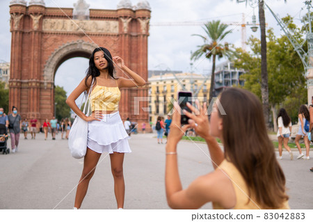 Young Asian female traveler in trendy summer outfit posing against triumphal arch while girlfriend taking photo on smartphone during sightseeing tour in Barcelona Young Asian female traveler in trendy summer outfit posing against triumphal arch while girlfriend taking photo on smartphone during sightseeing tour in Barcelona 83042883