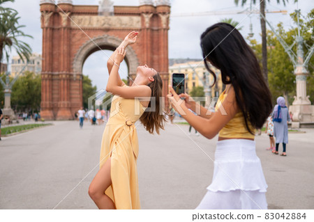Young woman in stylish yellow dress posing gracefully against historical triumphal arch while girlfriend taking picture on smartphone during summer holiday in Barcelona city Young woman in stylish yellow dress posing gracefully against historical triumphal arch while girlfriend taking picture on smartphone during summer holiday in Barcelona city 83042884