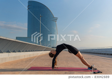 Side view of young woman in black activewear doing Purvottanasana pose against blue sky and skyscraper on city street. 83042889