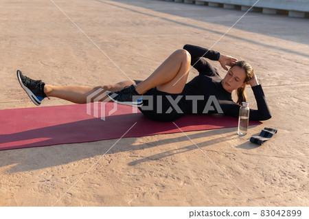 High angle of female athlete in sportswear and sneakers doing elbow to knee twists while lying on mat during fitness training on street. High angle of female athlete in sportswear and sneakers doing elbow to knee twists while lying on mat during fitness training on street. 83042899