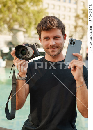 Choice between mobile phone and DSLR photo camera for photography. man holding two cameras while sitting near fountain on city square on sunny day 83042908