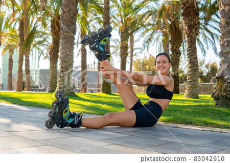Side view of happy female athlete in roller blades raising and stretching leg while sitting on pavement during workout in tropical park. 83042910
