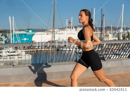 Focused sportswoman in sportswear running on embankment during cardio training in cloudless morning in harbor. 83042911