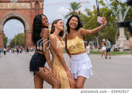 Stylish multiracial girlfriends in trendy clothes smiling brightly while taking selfie together on smartphone against triumphal arch of Barcelona city in summer day 83042927