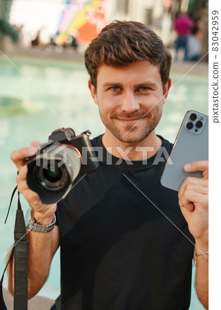 Happy young male photographer smiling and showing contemporary mobile phone and digital photo camera with optical lens while sitting near fountain in city park 83042959