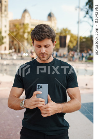 Concentrated young unshaven guy in casual clothes taking photo on camera of modern mobile phone while standing on city square on sunny day 83042961