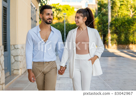 Trendy cheerful Hispanic couple holing hands and walking along street on summer day Trendy cheerful Hispanic couple holing hands and walking along street on summer day 83042967