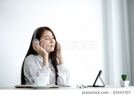 Video Calling a young Asian woman wearing headphones communicates via online long-distance video on a digital tablet happily greetings with a friend Video Calling a young Asian woman wearing headphones communicates via online long-distance video on a digital tablet happily greetings with a friend 83043707