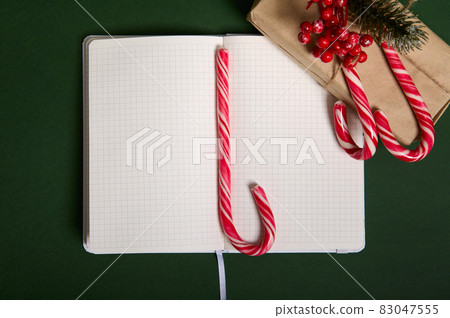 Christmas concepts. Beautiful simple present in craft wrapping gift paper with holly and candy canes on it, sugary striped lollipop on the middle of the notepad with blank empty pages with copy space Christmas concepts. Beautiful simple present in craft wrapping gift paper with holly and candy canes on it, sugary striped lollipop on the middle of the notepad with blank empty pages with copy space 83047555