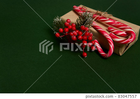 Christmas present in craft wrapping gift paper decorated with holly and sweet sugary striped white and red lollipops candy canes laid out in the corner of copy space on dark green background Christmas present in craft wrapping gift paper decorated with holly and sweet sugary striped white and red lollipops candy canes laid out in the corner of copy space on dark green background 83047557