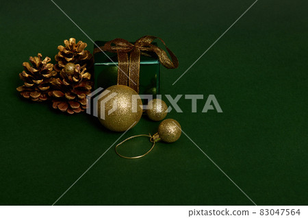 Luxury present in shiny green glitter wrapping gift paper with gold ribbon and golden bow, pines cones and spherical Christmas tree toys laid out on dark background. Copy space for advertisement 83047564