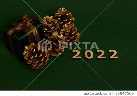 Beautiful composition of wooden numerals of the year 2022 next to luxury Christmas gift in shiny green gift wrapping paper and golden bow and pine cones colored in gold, isolated on dark backdrop 83047569
