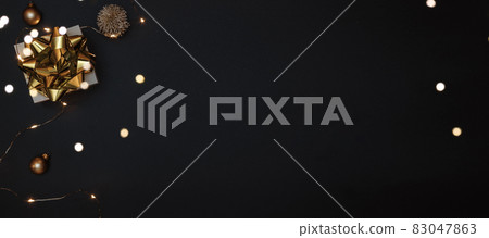 Christmas gift. White gift with golden bow, gold balls and sparkling lights garland in xmas decoration on dark background for greeting card. Xmas backdrop with space for text. Christmas gift. White gift with golden bow, gold balls and sparkling lights garland in xmas decoration on dark background for greeting card. Xmas backdrop with space for text. 83047863