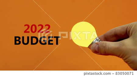 2022 budget new year symbol. Businessman holds yellow shining light bulb. Words '2022 budget', isolated on beautiful orange background. Business and 2022 budget new year concept. Copy space. 83050071