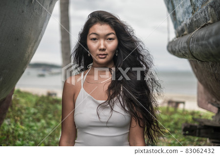 Portrait of beautiful young mongol woman in white dress standing between two old wooden ships. Looking at camera with copy space. Romantic portrait. Portrait of beautiful young mongol woman in white dress standing between two old wooden ships. Looking at camera with copy space. Romantic portrait. 83062432