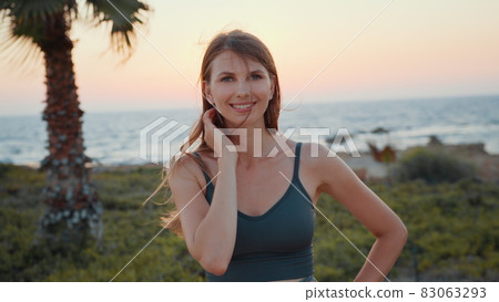 Portrait of caucasian woman in sport clothes smiling on camera while taking break after active workout on beach. Healthy and fit lady with long brown hair spending evening time for outdoors training. Portrait of caucasian woman in sport clothes smiling on camera while taking break after active workout on beach. Healthy and fit lady with long brown hair spending evening time for outdoors training. 83063293