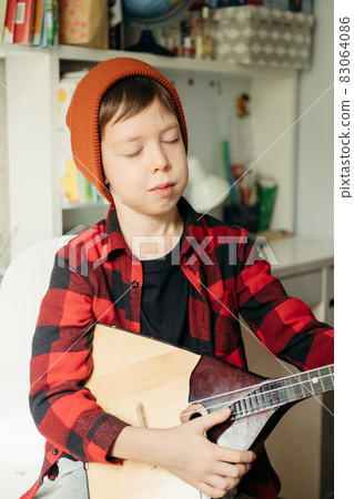 boy in a red hat and a plaid shirt plays the balalaika. Handsome boy holding his guitar. Music lessons at home. Hobby for the soul. home teaching music 83064086