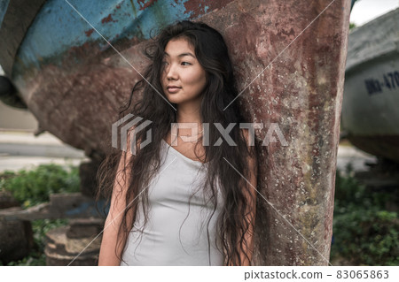 Portrait of beautiful young asian woman in white dress standing near the old ship and looking aside. Natural black curly long hair. Thinking face. 83065863