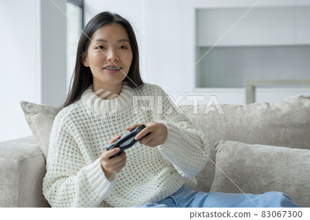 An Asian woman is sitting on the couch, holding a joystick and playing a game on the console. The woman looks excited with a joystick An Asian woman is sitting on the couch, holding a joystick and playing a game on the console. The woman looks excited with a joystick 83067300
