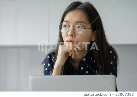 A thoughtful Asian businesswoman with glasses working with a laptop looks into the distance. The student uses virtual e-learning resources 83067398
