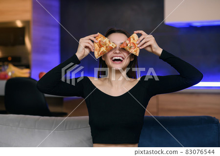 Excited happy young woman in kitchen plaing with pizza slices. Cover eyes with food. Carefree housekeeper very playful. Wear white dressing gown. Careless houseswife. Excited happy young woman in kitchen plaing with pizza slices. Cover eyes with food. Carefree housekeeper very playful. Wear white dressing gown. Careless houseswife. 83076544